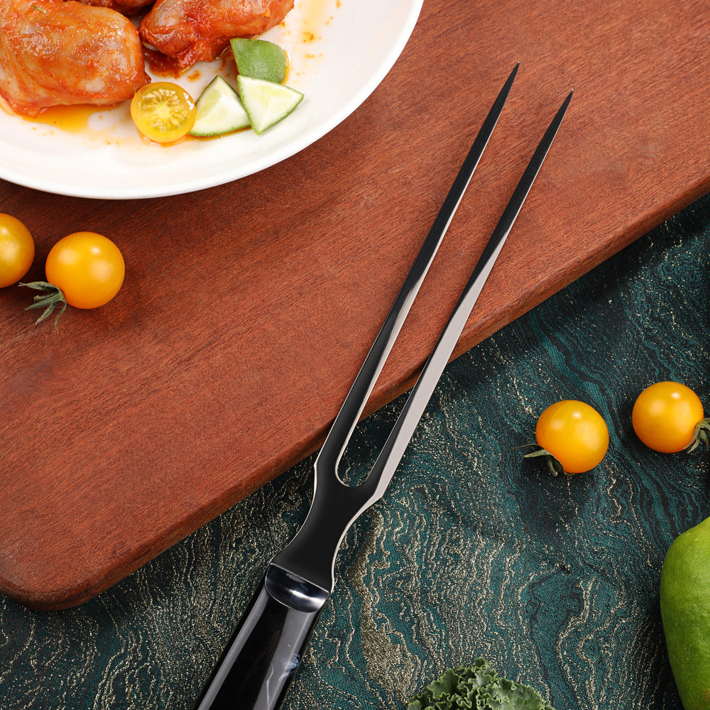 A black stainless steel two-prong carving fork with a textured handle, resting diagonally on a wooden cutting board surrounded by food props like cherry tomatoes, a lime, and cooked chicken.