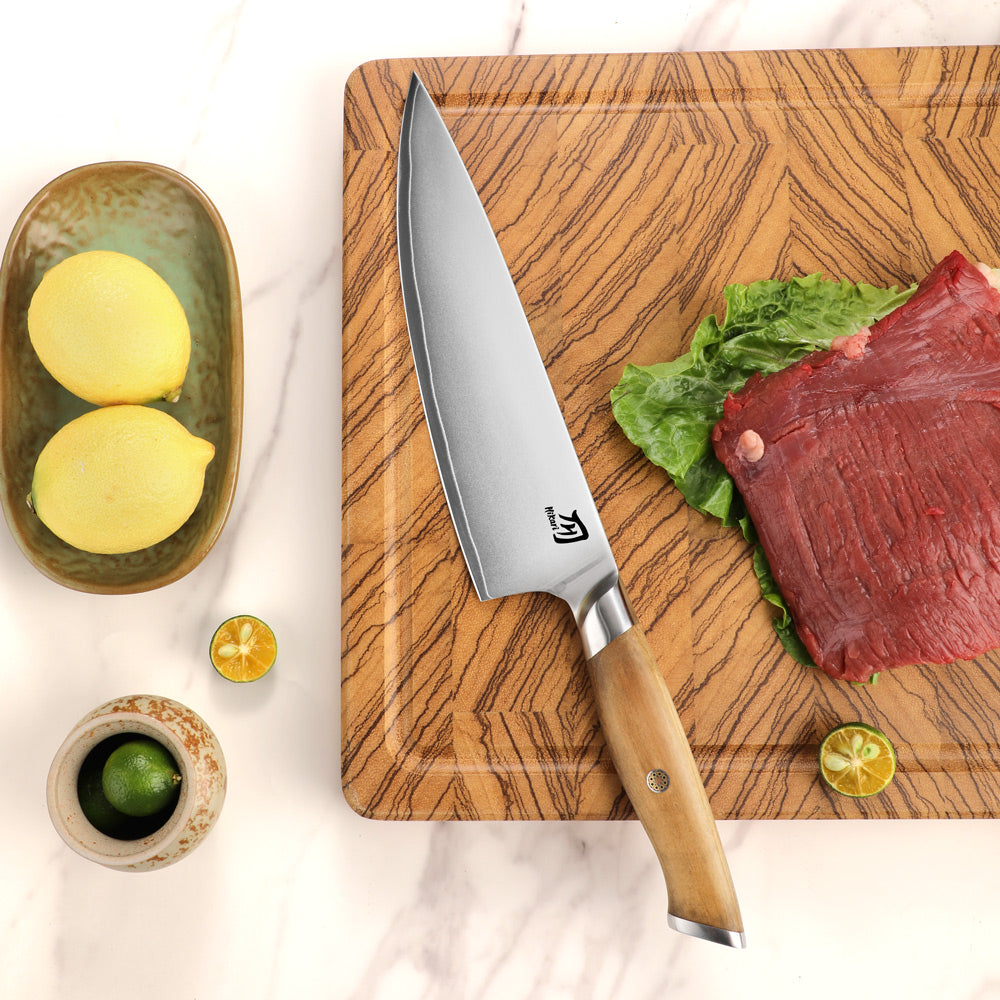 A chef's knife with a silver blade and light wood handle rests on a wooden cutting board next to raw beef, lettuce, lemons, and lime slices on a white marble surface.