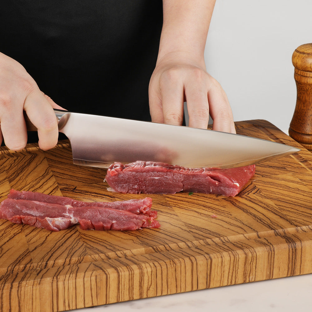 A close-up of a chef's knife slicing a piece of raw red meat on a thick wooden cutting board, with a person's hands and torso visible in a kitchen setting.