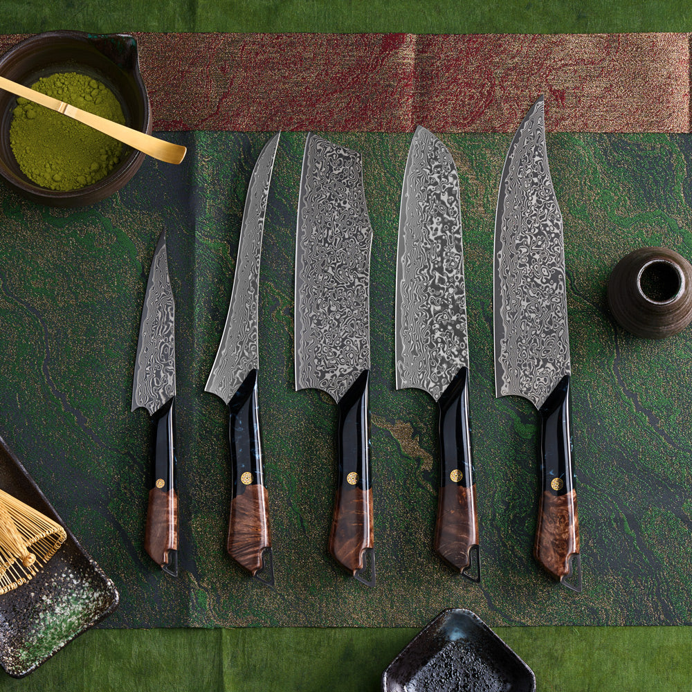 Five Japanese kitchen knives with patterned Damascus steel blades and dark wood handles are arranged on a green textured mat with traditional Japanese styling, including a bowl of green matcha, a bamboo whisk, and ceramic dishes.