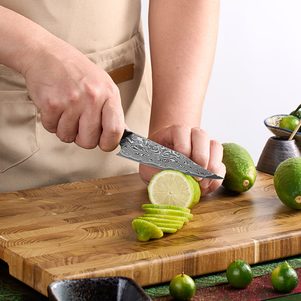 A close-up of a person slicing a lime on a wooden cutting board with a patterned steel knife. The person is wearing a beige apron, and several whole limes are visible around the cutting board.